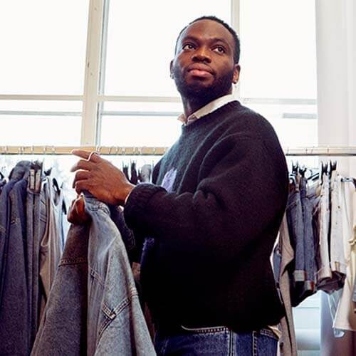 Un homme debout devant une tringle à vêtements. Il tient quelques vêtements et regarde hors caméra.